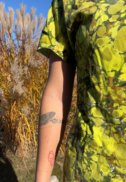 Person wearing a bright yellow patterned shirt with a tattoo on their arm, standing outdoors.